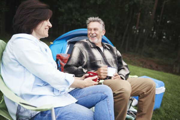 Älteres Paar sitzt in Campingstühlen neben dem Zelt und hält Tassen Tee, lächelnd