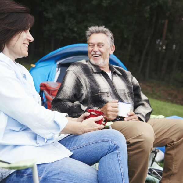 Mature couple sitting in camping chairs, beside tent, holding cups of tea, smiling