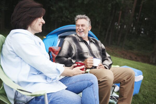 Mature couple sitting in camping chairs, beside tent, holding cups of tea, smiling